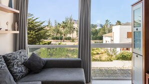 Un salón moderno con un sofá gris, un ventanal con cortinas y vistas a una zona residencial con árboles y edificios.