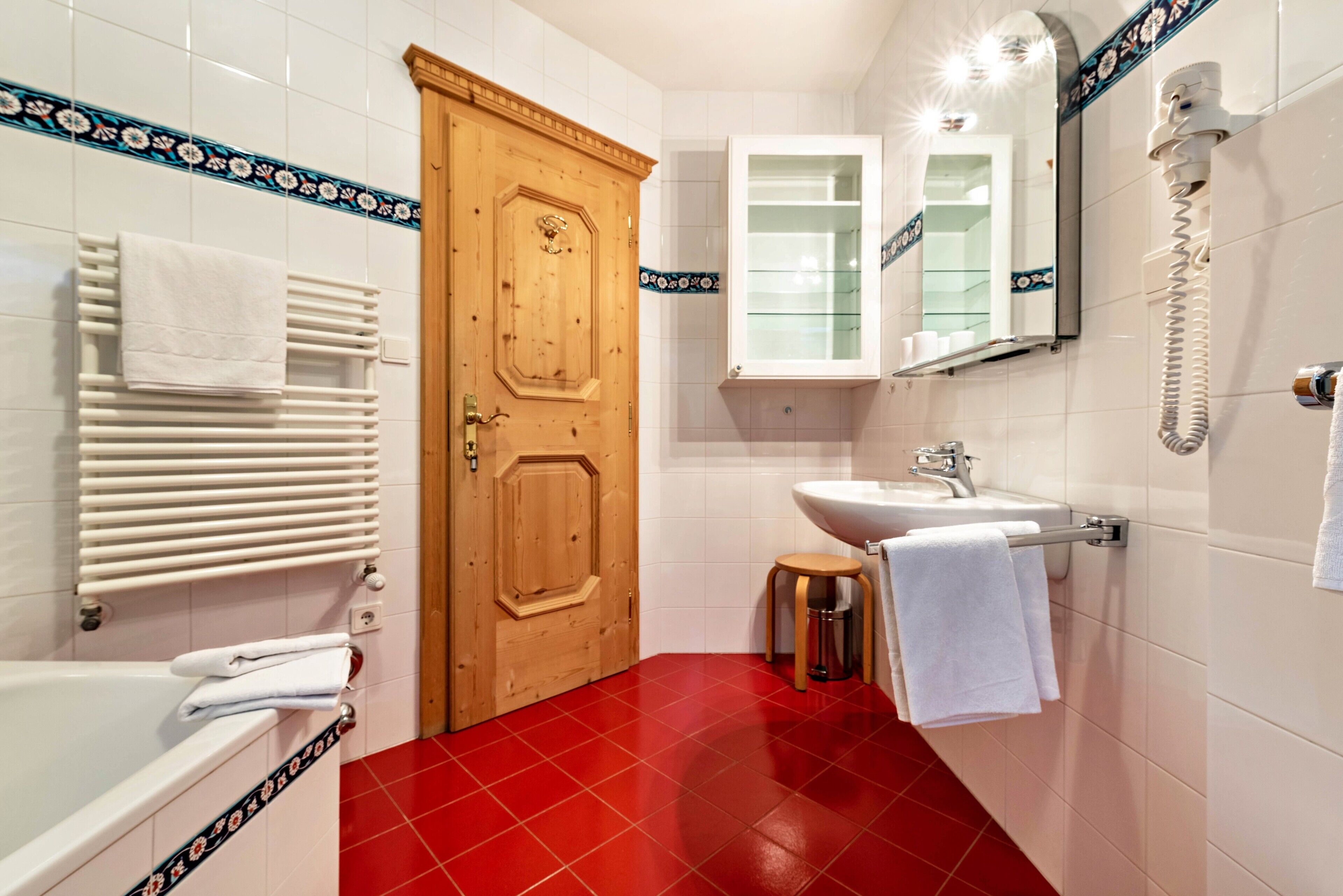 Un baño con puerta de madera, paredes de azulejos blancos, piso rojo, un lavamanos blanco y un espejo con teléfono.