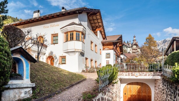 Un edificio tradizionale con una scala in pietra che conduce a una porta di legno, immerso nel verde sotto un cielo sereno.