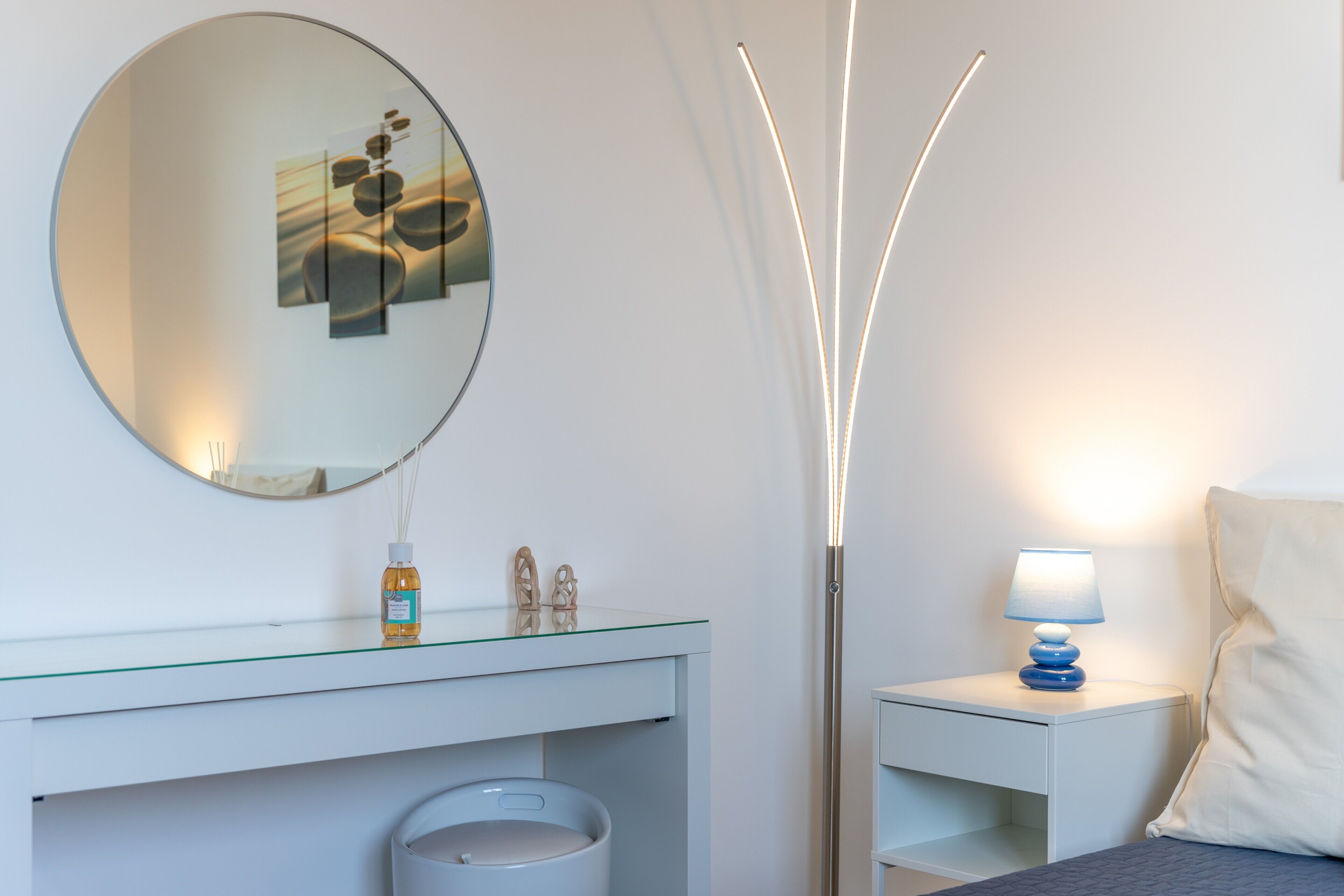 A modern bathroom with a round mirror, a glass-topped vanity, a white sink, a tall floor lamp, and a bedside table with a lamp.