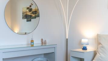 A modern bathroom with a round mirror, a glass-topped vanity, a white sink, a tall floor lamp, and a bedside table with a lamp.