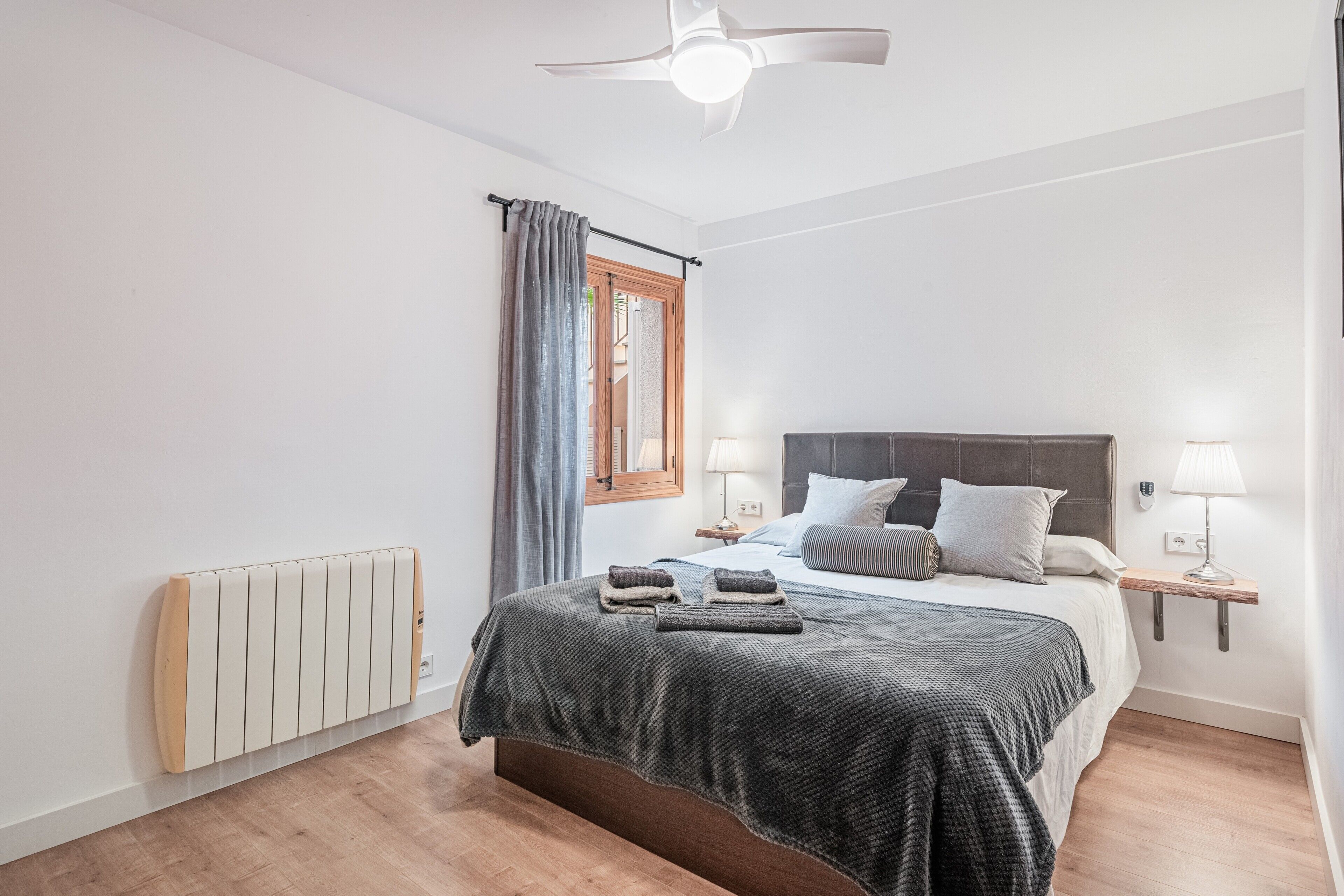 Un dormitorio con cama, lámparas de noche, ventilador de techo, ventana con cortinas y radiador.