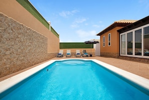 A swimming pool with lounge chairs, a stone wall, and a building with a tiled roof. - Vacation Home 'Villa Contenta' with Pool & Wi-Fi (Corralejo)