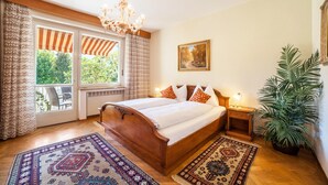 Een slaapkamer met een houten bed, een kroonluchter, een kamerplant en uitzicht op groen door het raam.