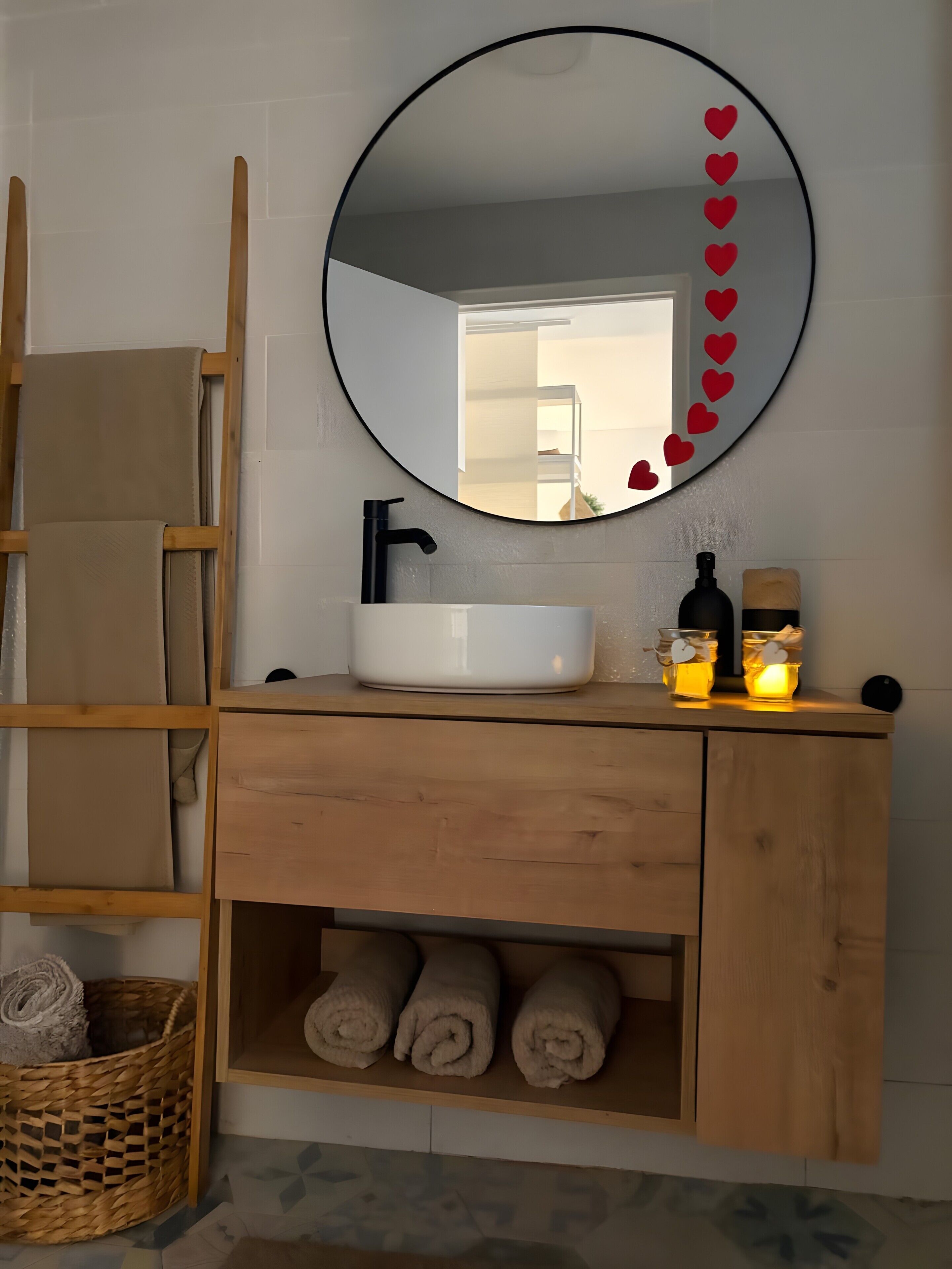 Een badkamer met een houten wastafelmeubel, een ronde spiegel met hartdecoraties, een witte wasbak en een zeepfles.