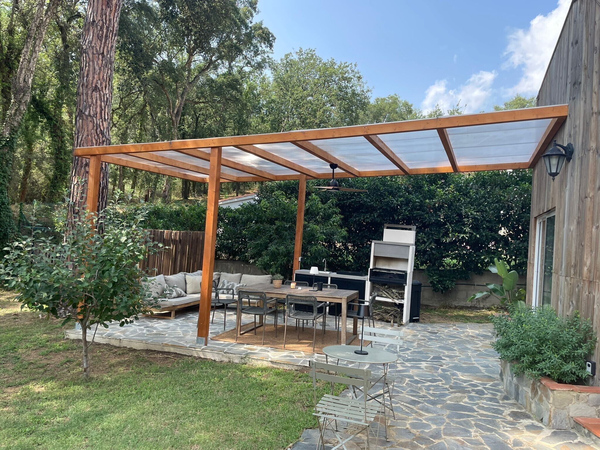 Een overdekt terras met stenen vloer, tuinmeubels en een barbecue.