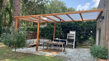 Een overdekt terras met stenen vloer, tuinmeubels en een barbecue.