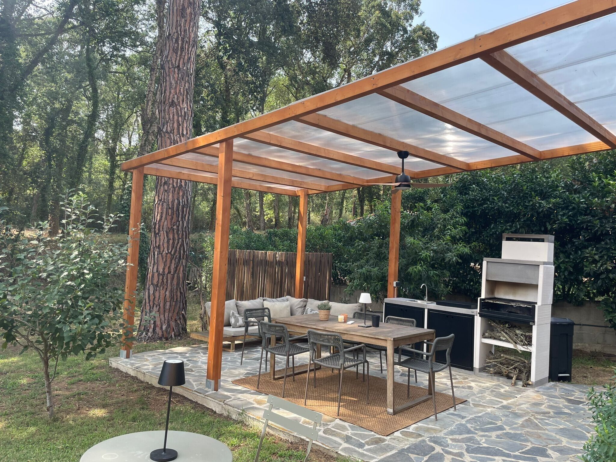 Eine überdachte Terrasse im Freien mit einer Holz-Pergola, Steinboden, einem Esstisch mit Stühlen, einem Grill und einer Sitzgruppe.