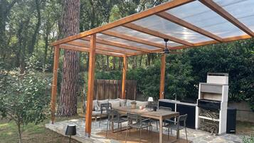 Eine überdachte Terrasse im Freien mit einer Holz-Pergola, Steinboden, einem Esstisch mit Stühlen, einem Grill und einer Sitzgruppe.