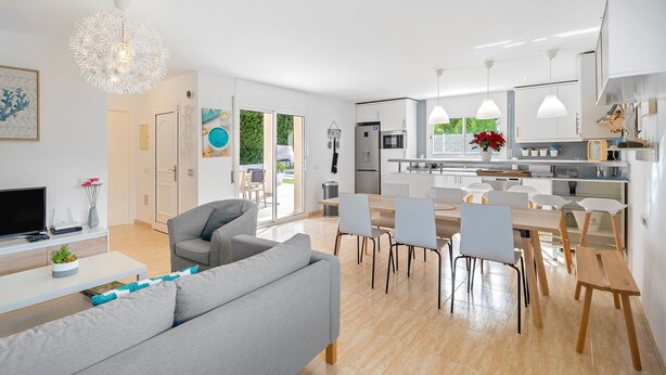 Une cuisine moderne avec des meubles blancs, un coin repas avec des chaises et un salon avec un canapé et une télévision.