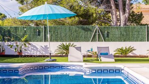 Une piscine dont le bord est revêtu de carreaux bleus et blancs, une haie verte et un parasol bleu.