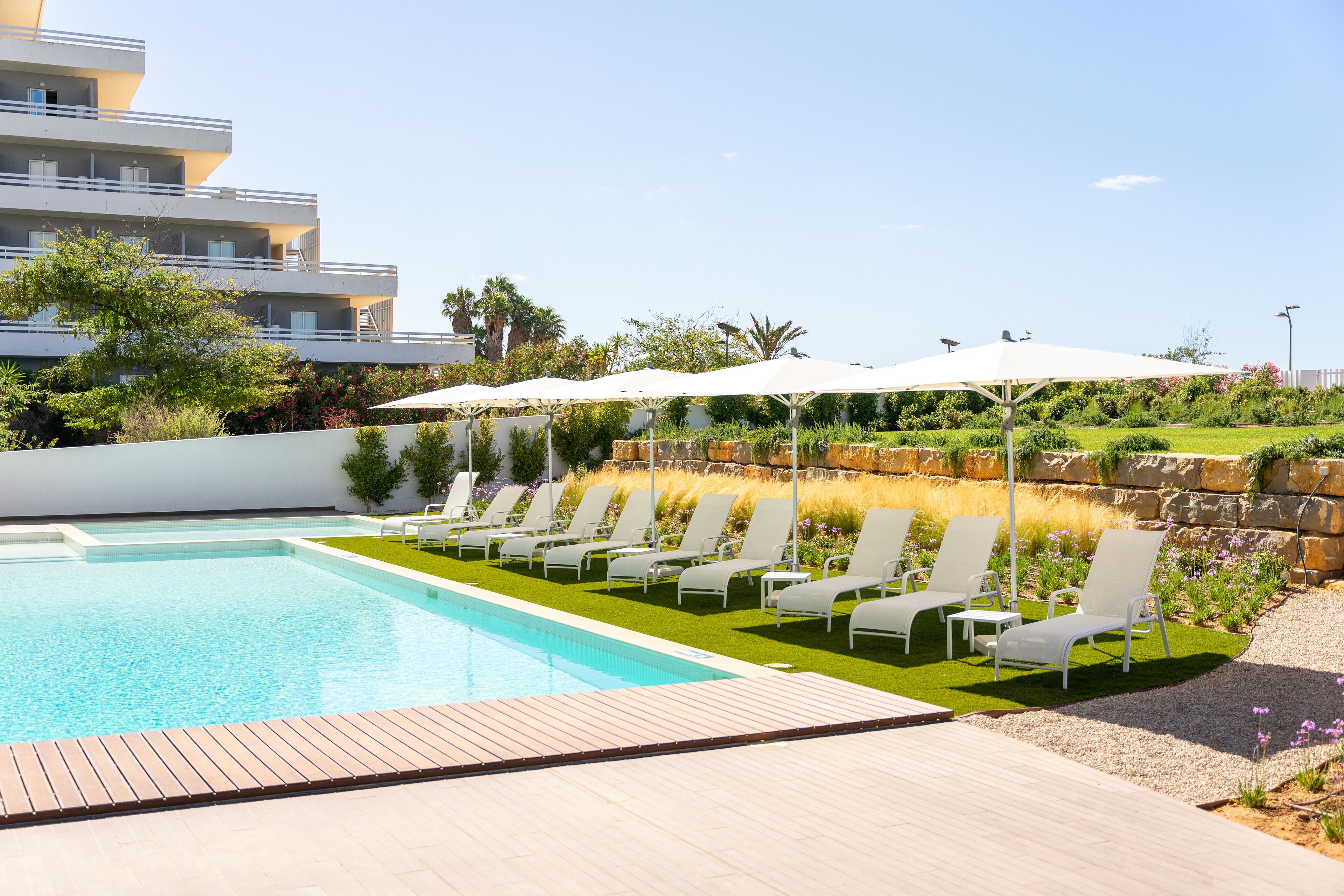 A swimming pool with lounge chairs and umbrellas, surrounded by greenery and a stone wall.