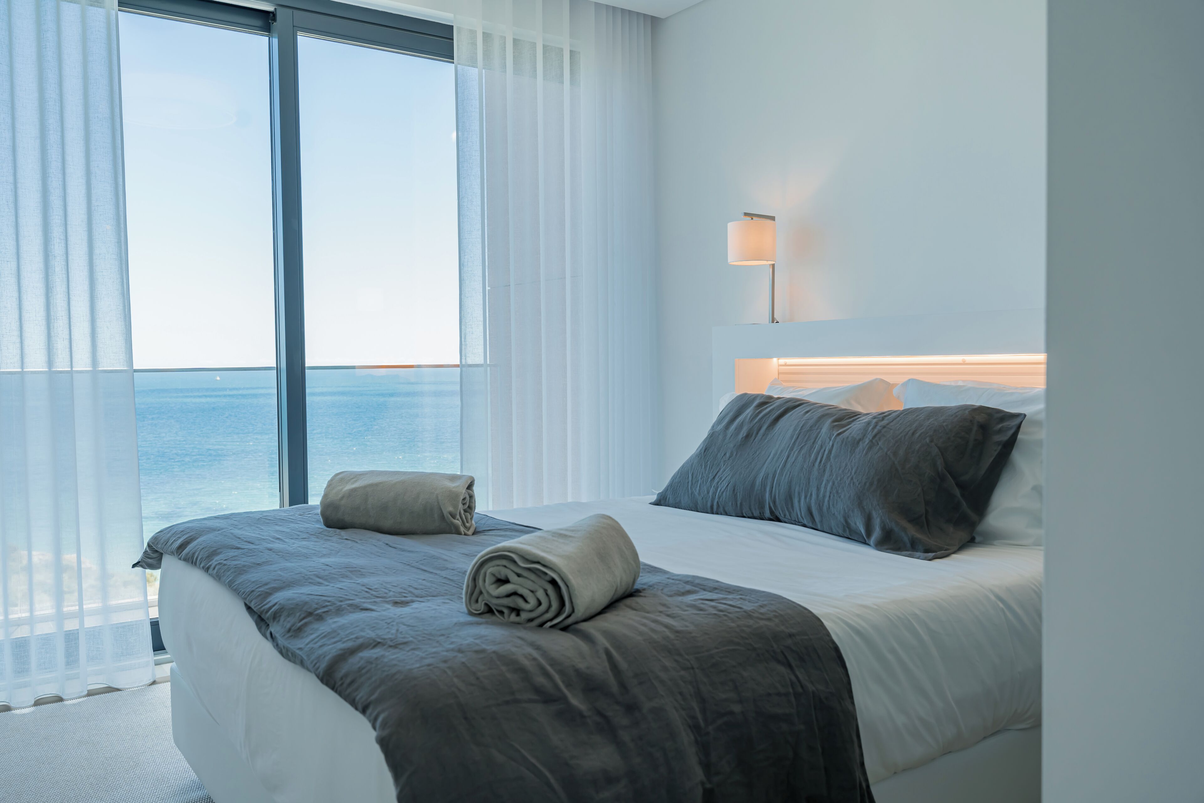 A modern hotel room with a large bed, a view of the sea, and a bedside lamp.