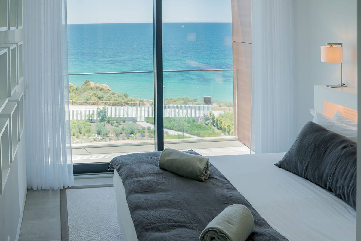 Ein Schlafzimmer mit großem Fenster und Meerblick, einem Bett mit grauer Bettwäsche sowie einem Nachttisch mit Lampe.