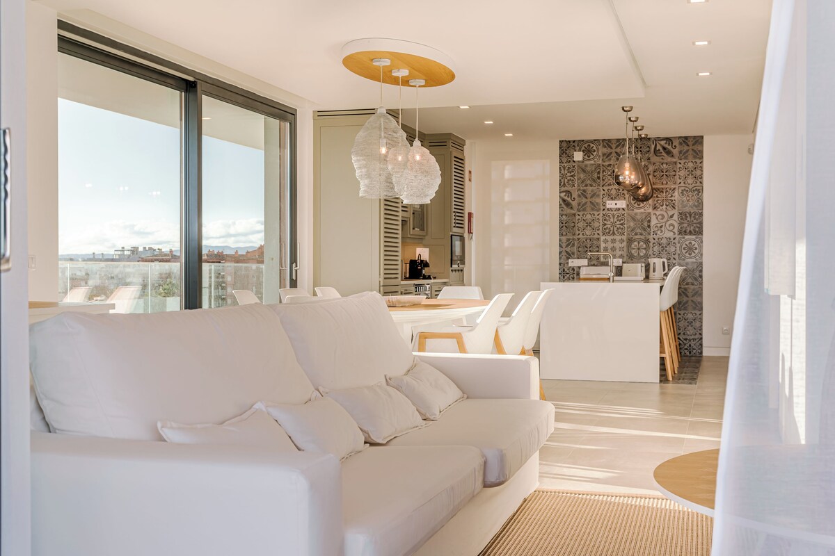 A modern living room with a white sofa, a dining area with a table and chairs, and a kitchen in the background.
