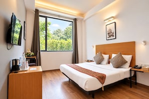 A hotel room with a bed, bedside table, lamp, TV, and a window with a view of greenery. - Enkay Residency Sector 15 (Gurugram)