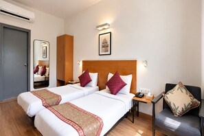 A hotel room with two beds, a wooden headboard, a bedside table with a phone and tissues, a chair with a floral cushion, and a framed picture on the wall. - Enkay Residency Sector 15 (Gurugram)