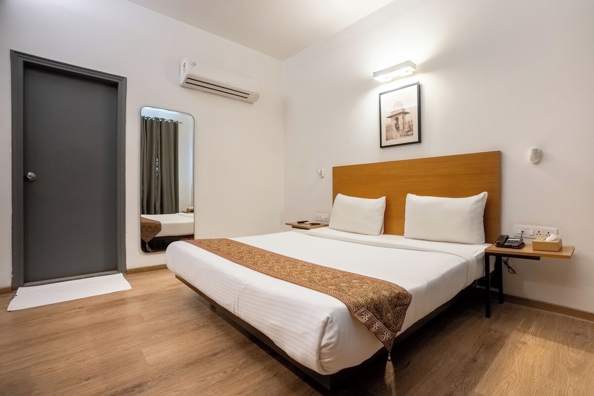A hotel room with a wooden bed, white bedding, a bedside table with a phone and tissues, a wall-mounted air conditioner, and a framed picture above the bed.