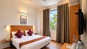 A hotel room with a bed, bedside table, a framed picture on the wall, a window with curtains, a television mounted on the wall, and a desk with a kettle, cups, and bottles. - Enkay Residency Sector 15 (Gurugram)