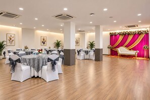 Un salón amplio con varias mesas redondas, sillas blancas con lazos grises y un escenario con cortinas doradas.
