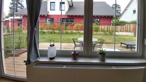 Una stanza con un'ampia finestra che si affaccia su un cortile con una casa rossa, un patio in legno e un tavolo con sedie.