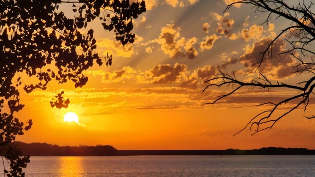 A sunset over a body of water with silhouettes of trees and a distant landmass. - Cozy private Lakehouse with prime fishing, hunting, and lake access. (Webber)