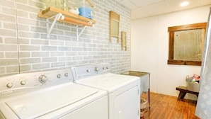 A laundry area with a brick wall, two white washing machines, and a wooden shelf with a jar and a blue container. - Cozy private Lakehouse with prime fishing, hunting, and lake access. (Webber)