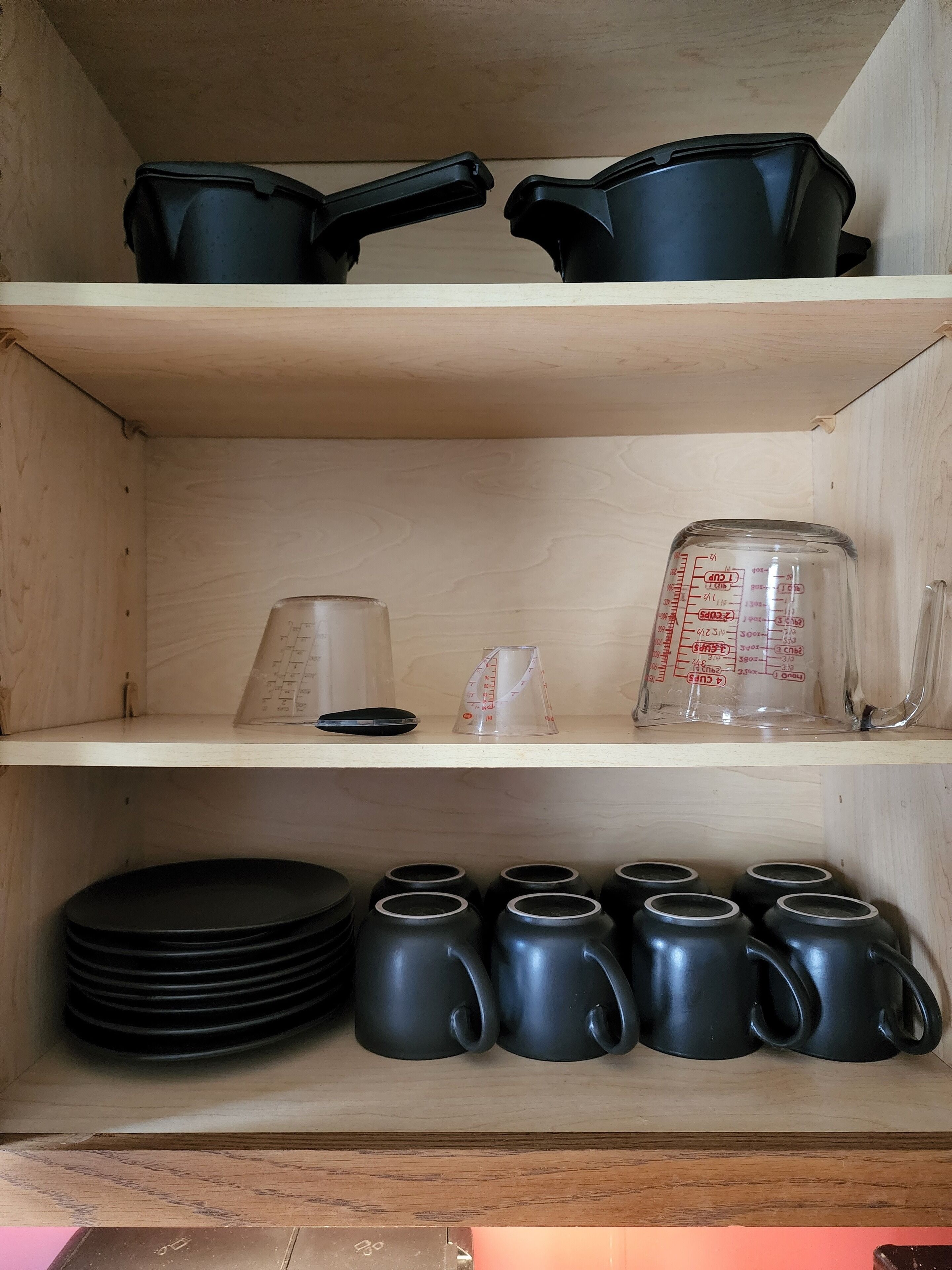 A kitchen shelf with black pots, a measuring cup, and stacked plates.