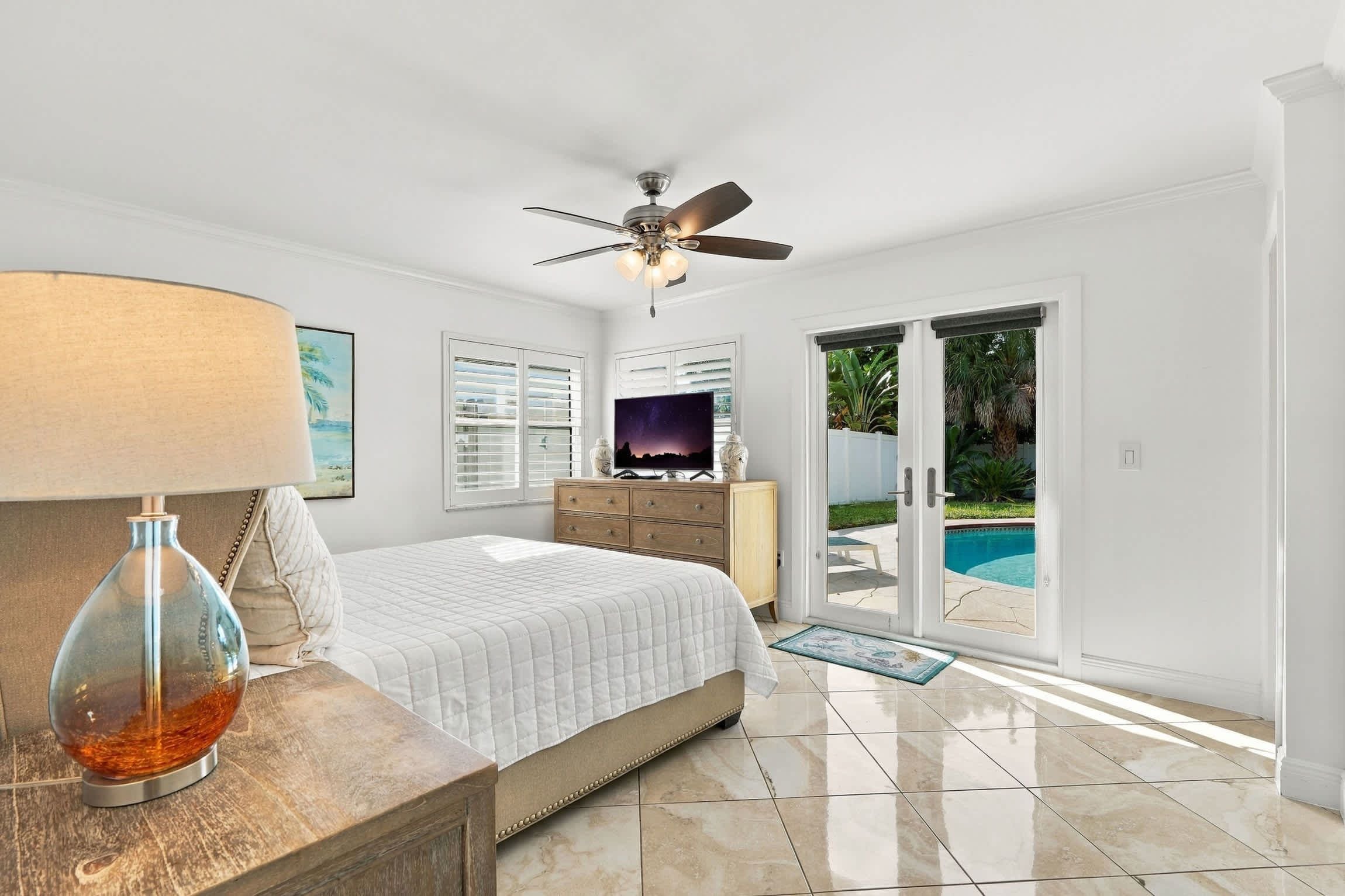 A bedroom with a bed, a ceiling fan, a TV, a wooden dresser, and a view of a swimming pool through a glass door.