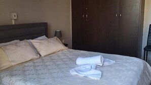 A neatly made bed with pillows and towels, a dark wooden wardrobe, and a small table with a lamp. - 2br-7 Guests-free Parking-pool-aircon (Paralimni)
