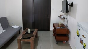 A small, well-lit room with a grey sofa, a wooden coffee table, a small table with a remote control, a wall-mounted TV, and an air conditioner. - 2br-7 Guests-free Parking-pool-aircon (Paralimni)