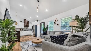A modern living room with a grey sofa, wooden coffee table, and a geometric rug. There is a kitchen area with white cabinets and a wooden accent wall.