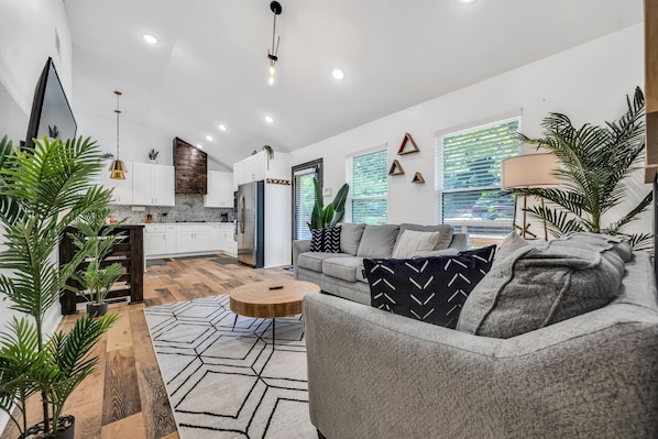 A modern living room with a grey sofa, wooden coffee table, and a geometric rug. There is a kitchen area with white cabinets and a wooden accent wall. - Harmony House, Sleeps 8 (Decatur)