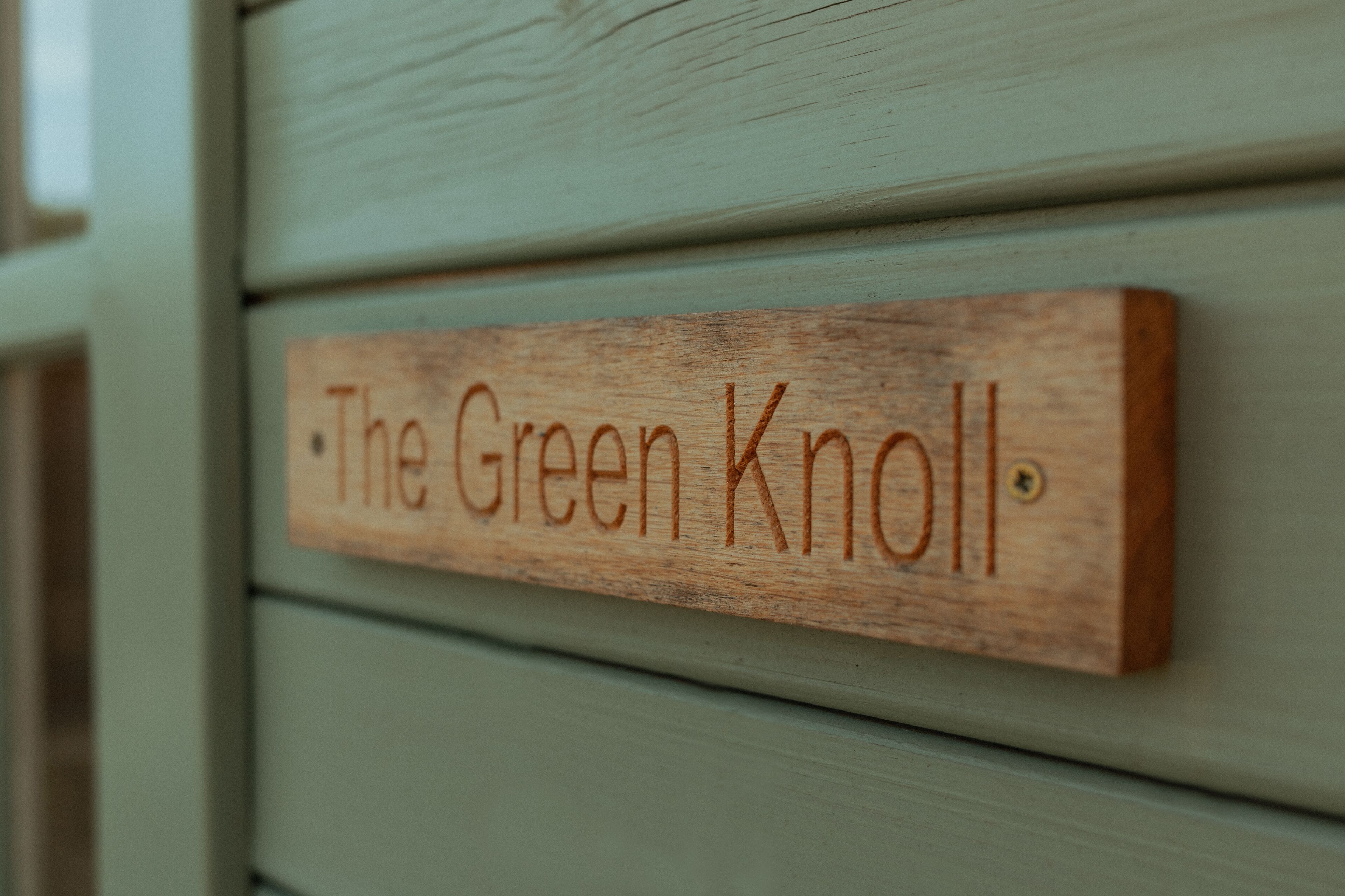 A wooden sign with the text "The Green Knoll" on a light green wall.