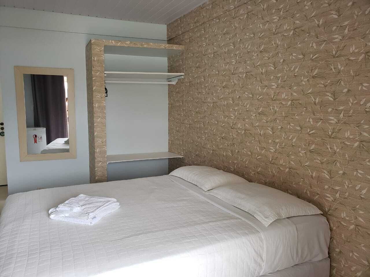 A neatly made bed with a white duvet and pillows, a folded towel on the bed, a mirror on the wall, and a shelf unit.