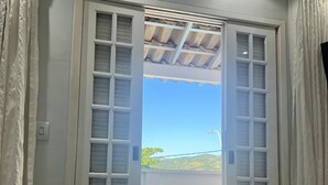 Une chambre avec un lit, une grande fenêtre donnant sur un quartier résidentiel et une porte donnant sur une rue.