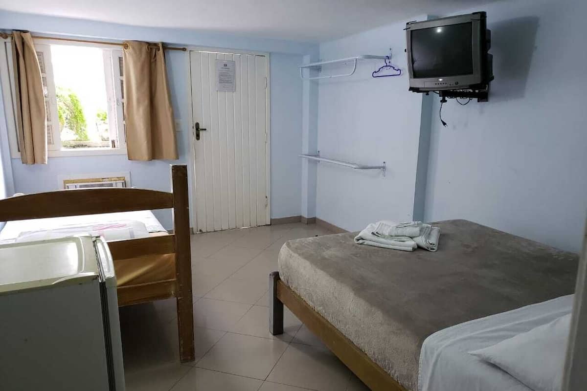 Ein Zimmer mit Bett, Stuhl, Schreibtisch, Fernseher und einem Fenster mit Vorhängen.