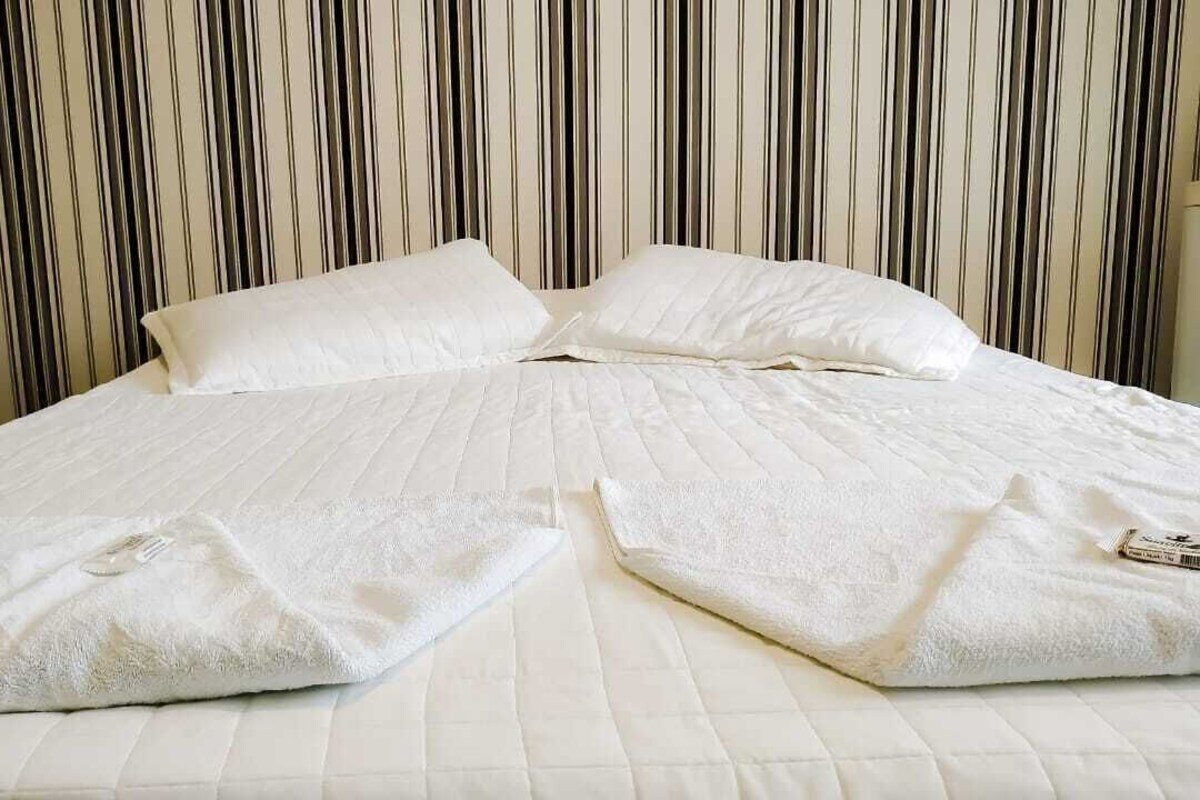 Una cama con ropa blanca y almohadas, un fondo de papel tapiz a rayas, y una mesita de noche con una lámpara.