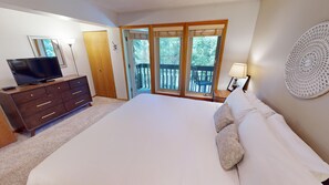 Ein Schlafzimmer mit einem großen Bett, einer Kommode, einem Fernseher und einem Balkon mit Blick auf Bäume.