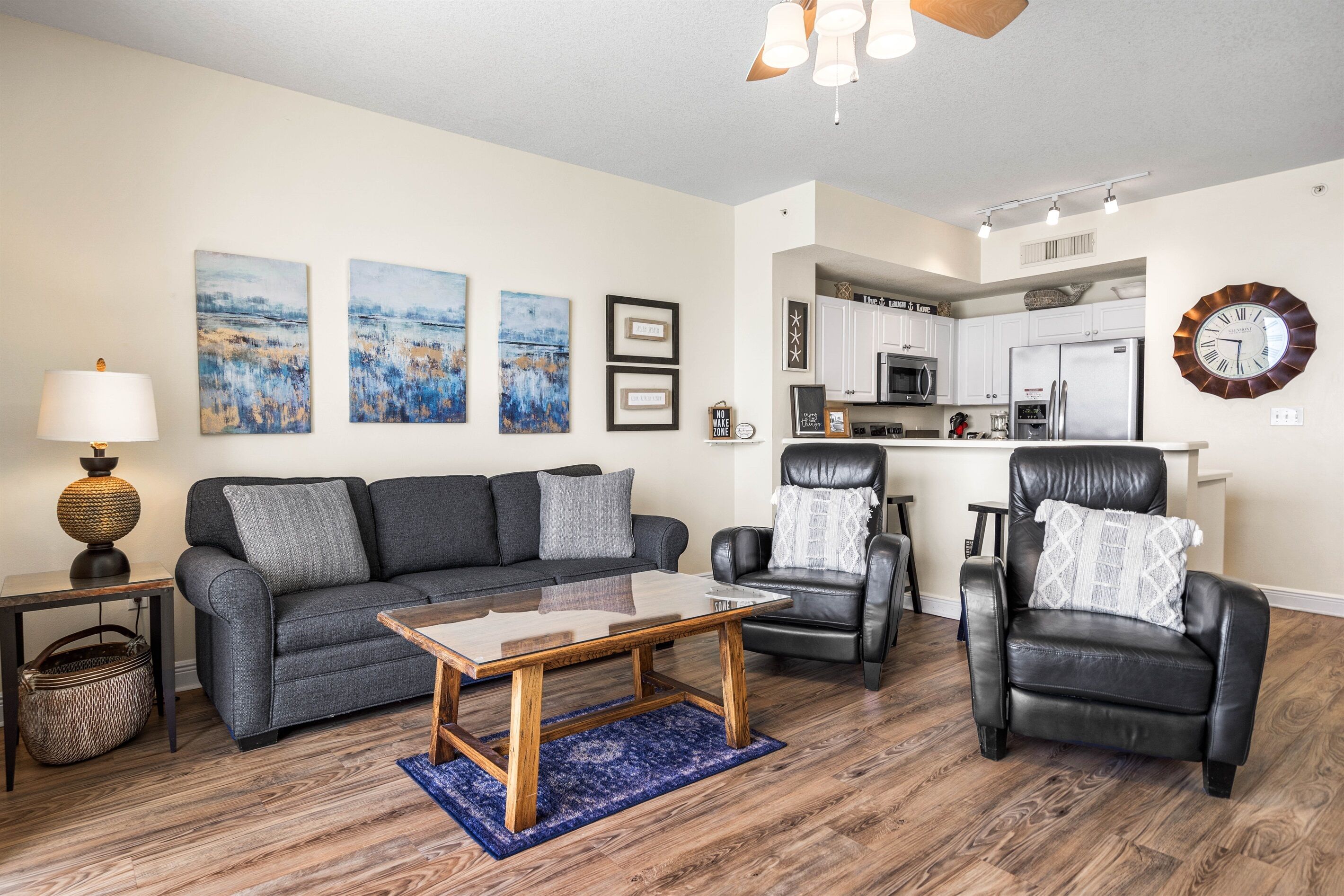 A living room with a sofa, coffee table, and two leather chairs. There is a kitchen in the background with white cabinets and stainless steel appliances.