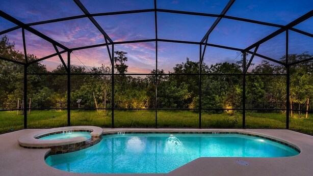 Une piscine au design incurvé, entourée d’une structure en verre et métal, se dressant sur un fond de verdure et sous un ciel crépusculaire.