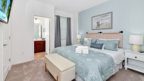Une chambre à coucher avec un lit, des tables de chevet, un banc, un téléviseur et une salle de bain visible à travers une porte ouverte.
