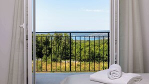 Ein Zimmer mit einem Bett, einem gerollten weißen Handtuch und Blick auf einen Balkon mit Pflanzen und einem Gewässer.