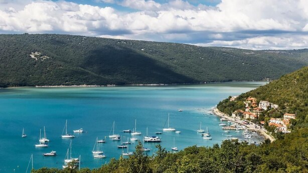 Eine Küstenstadt mit einer Marina voller Segelboote, umgeben von üppigen grünen Hügeln und wolkenlosem, blauem Himmel.