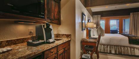 A hotel room with a kitchen area, a bed, a desk, and a window with curtains.