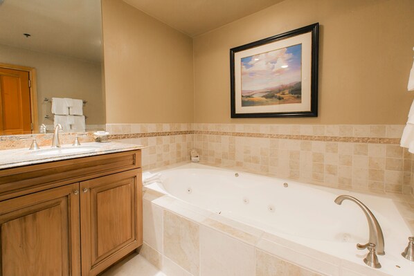 A bathroom with a large bathtub, wooden cabinets, a framed picture on the wall, and a tiled backsplash.