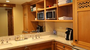 Baño de hotel con mueble de madera, encimera de mármol, lavamanos, dos microondas, cafetera y un perchero.