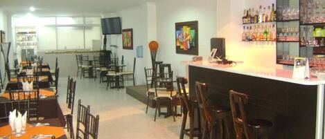 A restaurant with multiple dining tables and chairs, a bar area with a counter and shelves of bottles, and a television mounted on the wall.