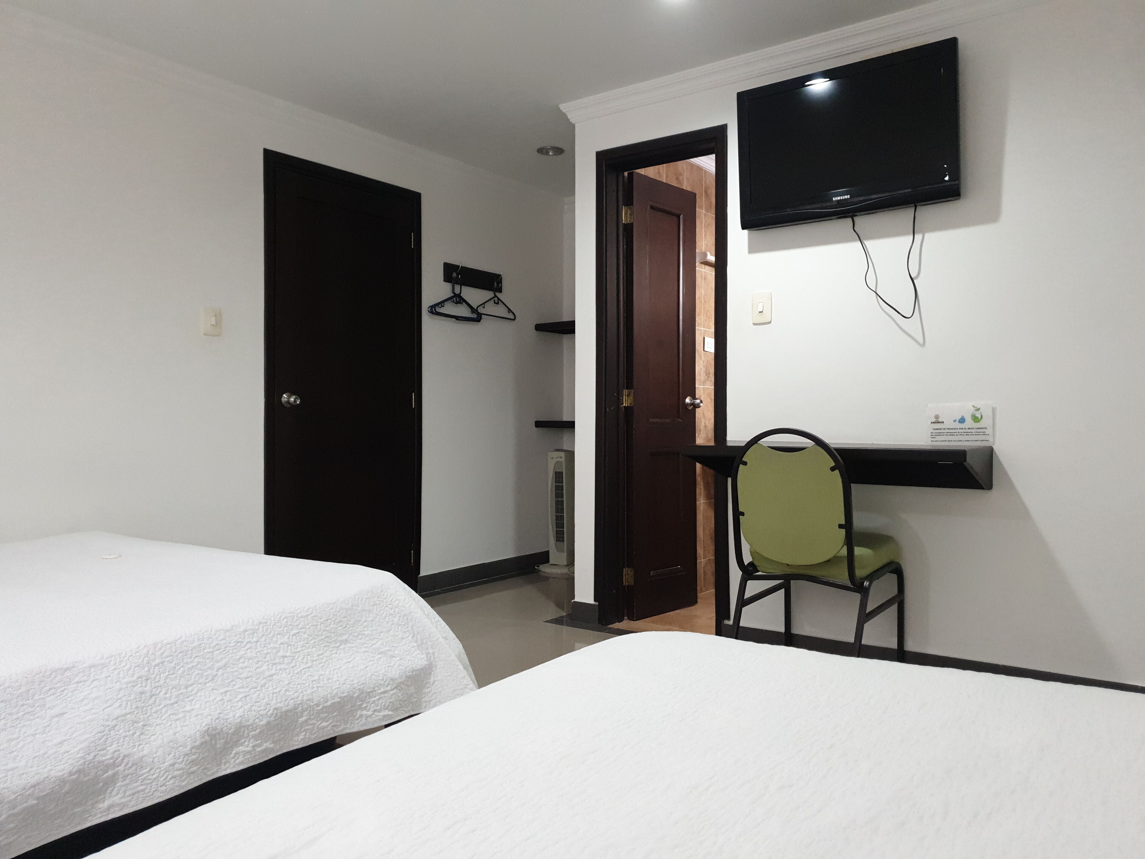 Une chambre d’hôtel avec deux lits simples, un téléviseur fixé au mur, une chaise et une porte donnant sur une autre pièce.