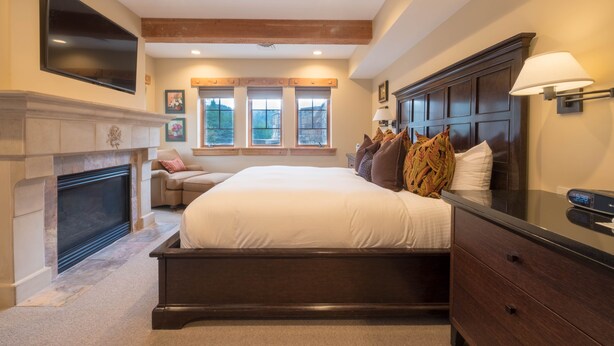 A bedroom with a large bed, a fireplace, a TV mounted on the wall, and a window with a view of trees.